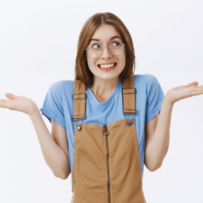 Waist-up shot of cute silly european female shrugging looking right with sorry smile spreading hands near shoulders being unaware and clueless acting like she knows nothing over gray background.
